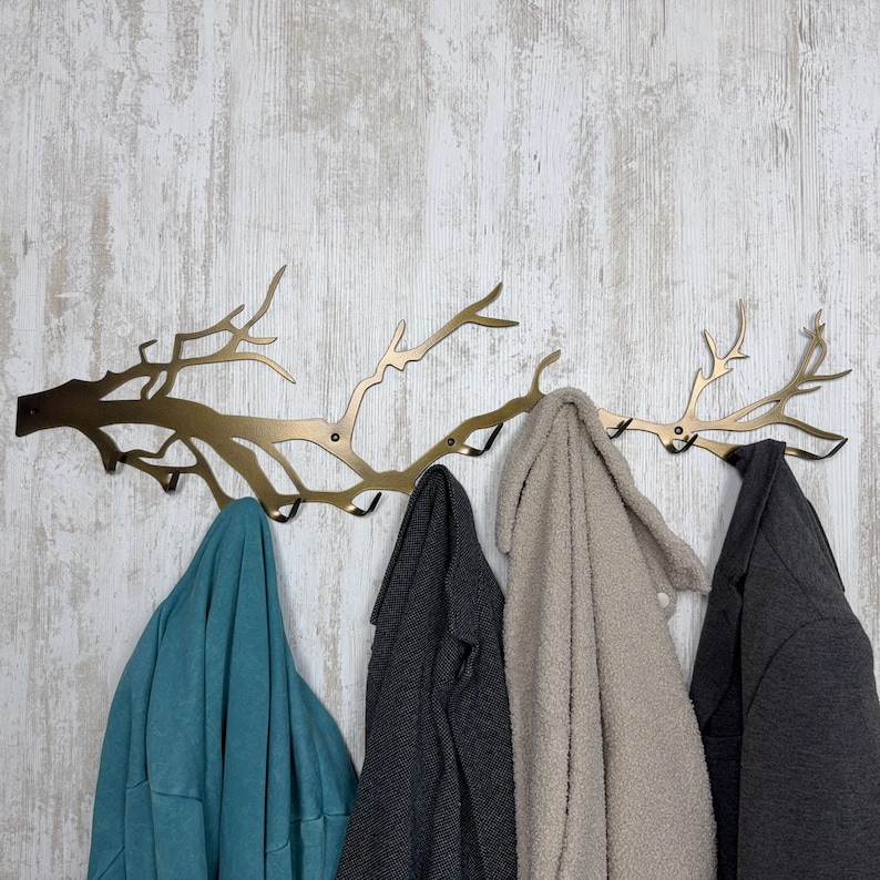 May include: A gold-colored metal wall-mounted coat rack in the shape of tree branches. Four coats in teal, gray, beige, and charcoal hang from the hooks. The rack is mounted on a white and gray wood-paneled wall.