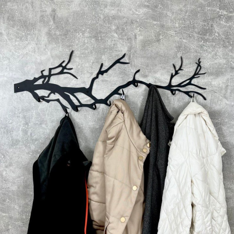 May include: A black metal wall-mounted coat rack shaped like a tree branch, with multiple hooks. Four coats in black, tan, gray, and white hang from the hooks. The rack is mounted on a gray textured wall.