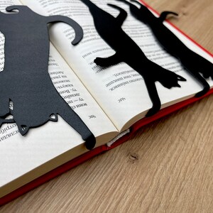 Pu&ograve; includere: Tre segnalibri di metallo nero a forma di gatto posizionati su un libro aperto con una copertina rossa. I segnalibri hanno la forma di un gatto in diverse pose.