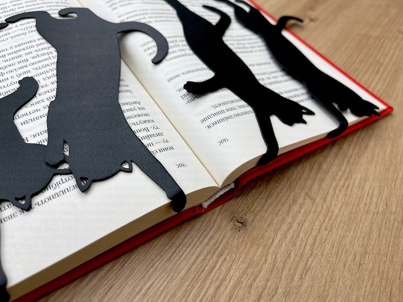 Pu&ograve; includere: Tre segnalibri di metallo nero a forma di gatto posizionati su un libro aperto con una copertina rossa. I segnalibri hanno la forma di un gatto in diverse pose.