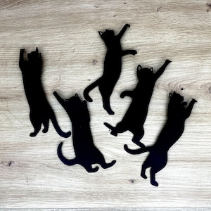 Pu&ograve; includere: Cinque sagome di gatti neri sono disposte su una superficie di legno marrone chiaro. I gatti sono in varie pose giocose, con le code arrotolate e i corpi allungati.