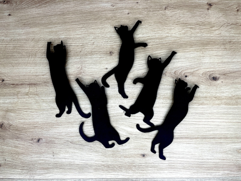 Pu&ograve; includere: Cinque sagome di gatti neri sono disposte su una superficie di legno marrone chiaro. I gatti sono in varie pose giocose, con le code arrotolate e i corpi allungati.