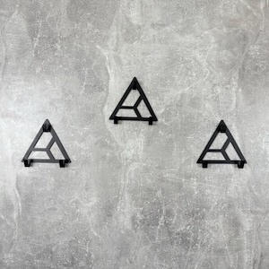 以下が含まれることがあります： グレーのテクスチャード加工された壁に、黒い金属製の三角形のウォールフックが3つあります。各フックは、下部に小さな棚が付いた幾何学的なデザインです。フックは均等に配置されています。