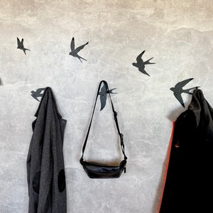May include: A wall with five black bird-shaped hooks, two of which are holding a gray sweater and a black jacket. A black leather bag hangs from one of the hooks.