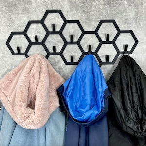 May include: Black metal wall-mounted coat rack with a honeycomb design. The rack holds three coats: a light blue denim jacket with a pink faux fur hood, a navy blue jacket with a blue interior, and a black jacket.