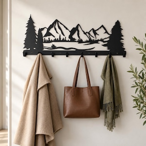 May include: A black metal wall-mounted coat rack featuring a laser-cut silhouette of a mountain range flanked by two pine trees. The horizontal rack includes seven evenly spaced hooks along the bottom edge for hanging various items.