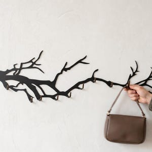 May include: A black metal wall-mounted coat rack in the shape of a tree branch, with multiple hooks. A brown purse is hanging from one of the hooks. The rack is mounted on a white wall.