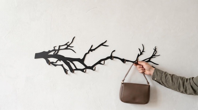 May include: A black metal wall-mounted coat rack in the shape of a tree branch, with multiple hooks. A brown purse is hanging from one of the hooks. The rack is mounted on a white wall.