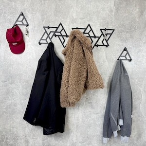 以下が含まれることがあります： 幾何学模様のモダンな壁掛けコートラック。バーガンディの野球帽と鍵が別のフックに掛かっています。黒いジャケット、茶色のふわふわのコート、グレーのパーカーがラックに掛けられています。壁は明るいグレーです。