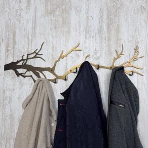 May include: A decorative wall-mounted coat rack in the shape of a tree branch, made of gold-colored metal. Three coats hang from the rack: a beige fleece coat, a navy blue quilted jacket, and a gray tweed coat.