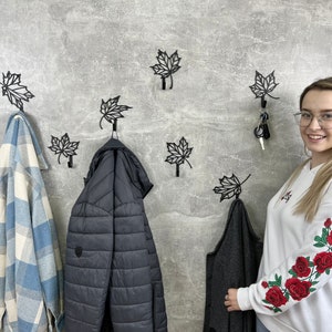 May include: A wall with black metal leaf-shaped hooks. There are two jackets hanging on the hooks, one is blue and white plaid and the other is black. A woman is standing to the right of the hooks, wearing a white sweatshirt with red roses embroidered on the sleeves.