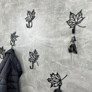 May include: Six black metal leaf-shaped hooks mounted on a gray wall. Two hooks have items hanging from them, including a black jacket and a blue and white plaid blanket. One hook has a set of keys hanging from it.