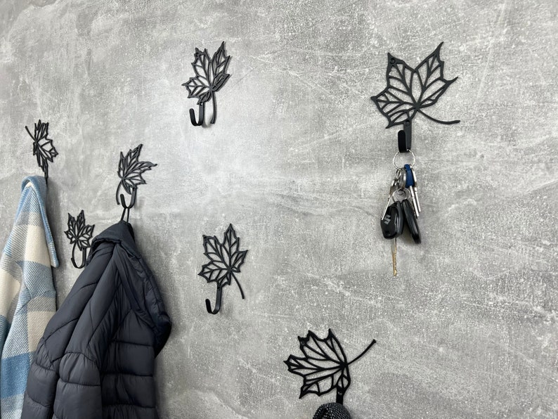 May include: Six black metal leaf-shaped hooks mounted on a gray wall. Two hooks have items hanging from them, including a black jacket and a blue and white plaid blanket. One hook has a set of keys hanging from it.