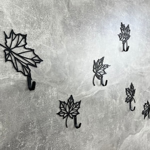 May include: Seven black metal hooks shaped like maple leaves mounted on a gray wall. Each hook has a single point at the bottom for hanging items.