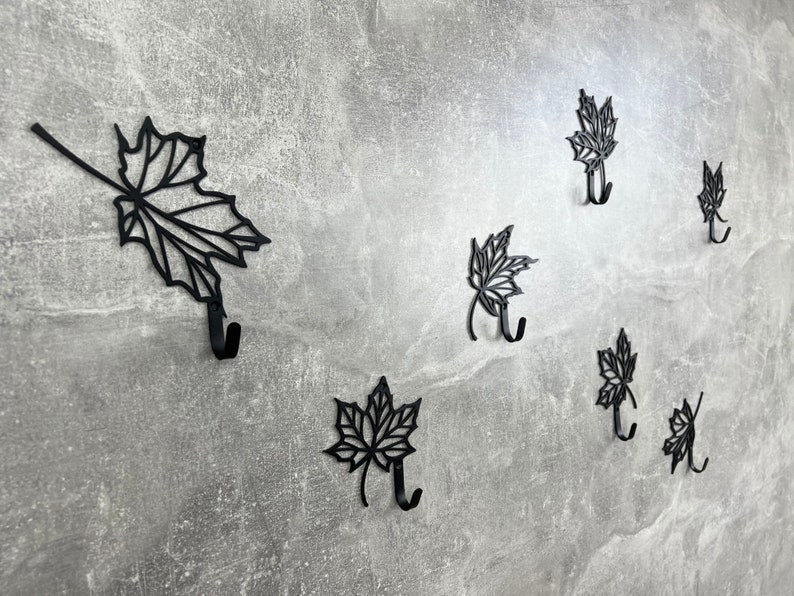 May include: Seven black metal hooks shaped like maple leaves mounted on a gray wall. Each hook has a single point at the bottom for hanging items.