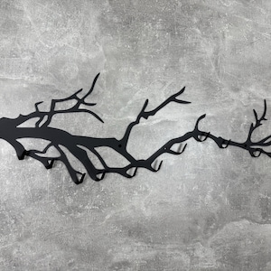 May include: Black metal wall-mounted coat rack shaped like a tree branch with multiple hooks. The rack is mounted on a gray textured wall. The design is a decorative and functional storage solution.