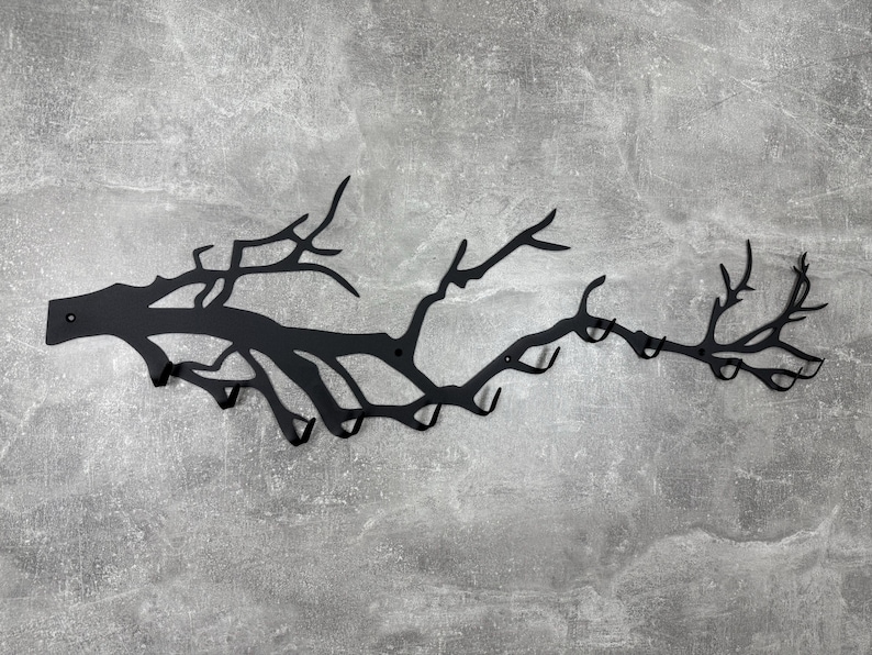 May include: Black metal wall-mounted coat rack shaped like a tree branch with multiple hooks. The rack is mounted on a gray textured wall. The design is a decorative and functional storage solution.