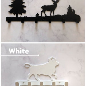 Puede incluir: Porta llaves de metal negro y blanco con una silueta de ciervo y una escena de bosque. La versi&oacute;n blanca tiene una silueta de toro y se muestra con llaves colgando de los ganchos.
