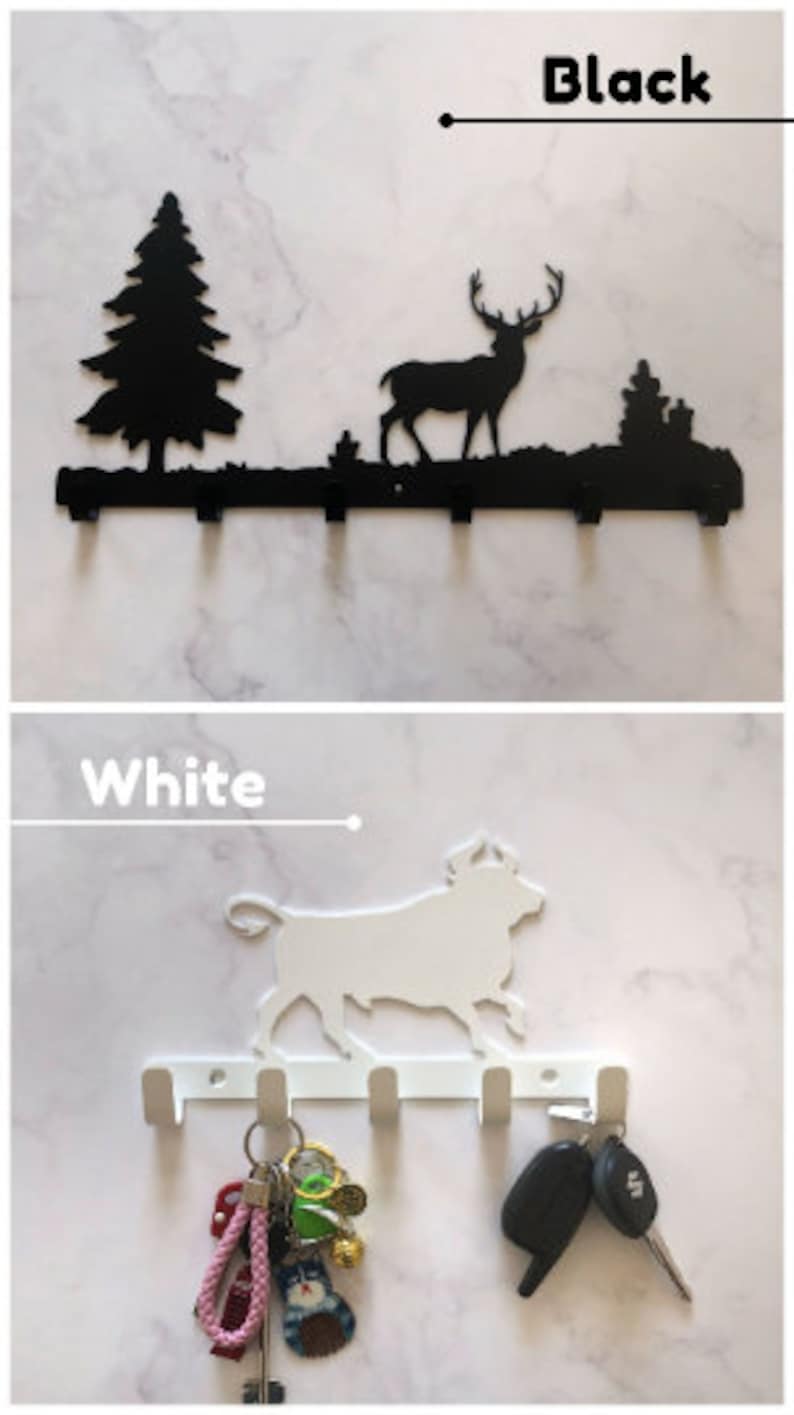 Puede incluir: Porta llaves de metal negro y blanco con una silueta de ciervo y una escena de bosque. La versi&oacute;n blanca tiene una silueta de toro y se muestra con llaves colgando de los ganchos.