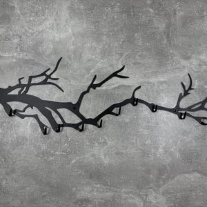 May include: Black metal wall-mounted coat rack in the shape of a tree branch. The rack has multiple hooks for hanging coats, hats, or other items. The design is a silhouette of a tree branch with a matte finish.