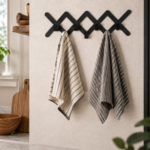 Könnte beinhalten: Eine ausziehbare Wandgarderobe aus schwarzem Metall mit fünf Haken und einem kreuzförmigen Design. An den Haken hängen zwei Küchentücher: eines ist cremefarben mit dünnen schwarzen Streifen, das andere ist dunkelgrau mit weißen Streifen.