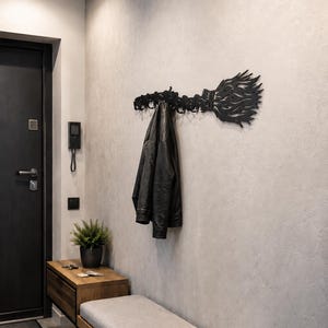 May include: A black metal wall-mounted coat rack shaped like a stylized broomstick with multiple hooks. The rack is mounted on a light gray wall and holds a black leather jacket. The design features intricate, flame-like patterns on the broom head.