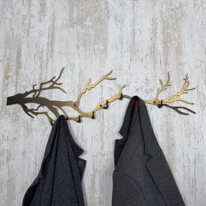 May include: A decorative metal wall-mounted coat rack in the shape of a tree branch. The rack is a mix of bronze and gold tones, with multiple hooks for hanging coats. Two gray coats are hanging on the rack against a light wood-grain background.