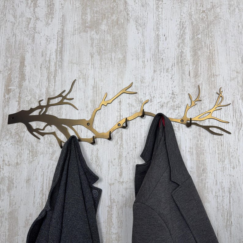 May include: A decorative metal wall-mounted coat rack in the shape of a tree branch. The rack is a mix of bronze and gold tones, with multiple hooks for hanging coats. Two gray coats are hanging on the rack against a light wood-grain background.