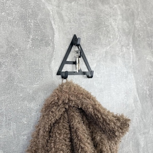 Può includere: Un portachiavi e attaccapanni da parete in metallo nero a forma triangolare. Il portachiavi contiene chiavi e un cappotto marrone e soffice. L'attaccapanni è montato su un muro grigio strutturato.