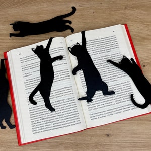 Pu&ograve; includere: Cinque segnalibri neri a forma di gatto sono posizionati su un libro aperto. I segnalibri sono realizzati in un materiale nero sottile e hanno la forma di gatti in varie pose. Il libro &egrave; aperto a una pagina con testo in una lingua straniera.