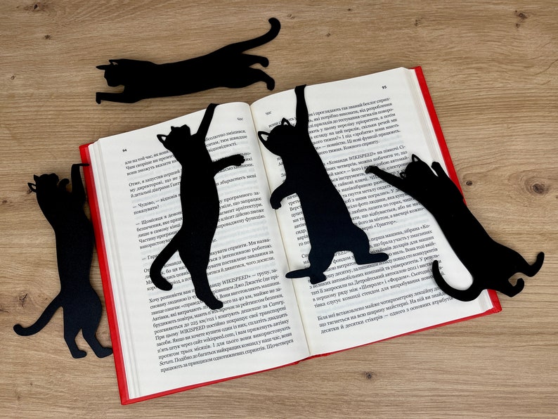 Pu&ograve; includere: Cinque segnalibri neri a forma di gatto sono posizionati su un libro aperto. I segnalibri sono realizzati in un materiale nero sottile e hanno la forma di gatti in varie pose. Il libro &egrave; aperto a una pagina con testo in una lingua straniera.