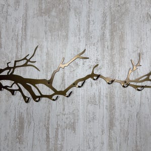 May include: A bronze-colored metal wall-mounted coat rack shaped like a tree branch. The rack has multiple hooks for hanging items and is mounted on a light-colored, wood-look wall.