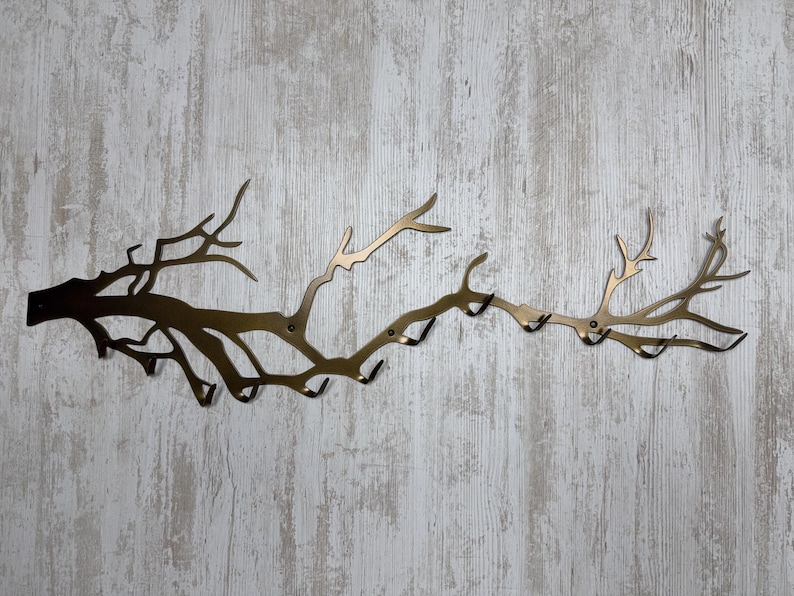 May include: A bronze-colored metal wall-mounted coat rack shaped like a tree branch. The rack has multiple hooks for hanging items and is mounted on a light-colored, wood-look wall.