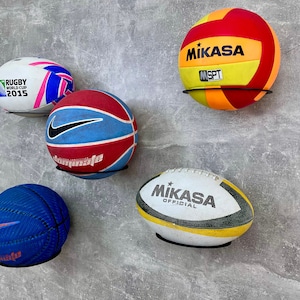 Puede incluir: Cuatro balones de diferentes deportes montados en una pared. Una pelota de rugby blanca y rosa, una pelota de baloncesto azul y roja, una pelota de voleibol amarilla y roja con el texto "MIKASA" y una pelota de rugby blanca y amarilla con el texto "MIKASA OFFICIAL".