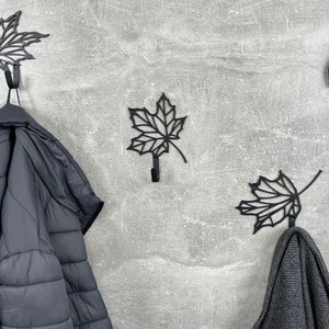 May include: Four black metal leaf-shaped hooks mounted on a gray wall. Two hooks have black jackets hanging from them.