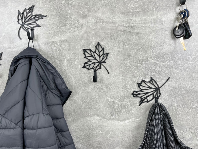 May include: Four black metal leaf-shaped hooks mounted on a gray wall. Two hooks have black jackets hanging from them.