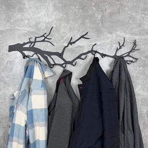 May include: A dark gray metal wall-mounted coat rack shaped like tree branches, with multiple hooks. Various coats and jackets hang from the hooks, including a blue and white plaid shirt, a gray hoodie, a navy blue quilted jacket, and a gray wool coat.