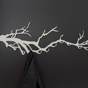 May include: A white metal wall-mounted coat rack shaped like a tree branch. The rack has multiple hooks for hanging items and is mounted on a dark gray wall. A dark gray jacket hangs from one of the hooks.