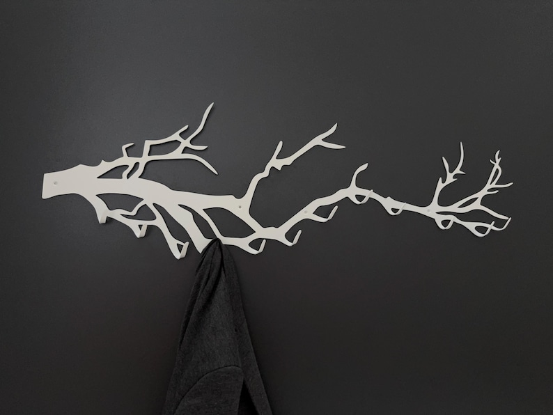 May include: A white metal wall-mounted coat rack shaped like a tree branch. The rack has multiple hooks for hanging items and is mounted on a dark gray wall. A dark gray jacket hangs from one of the hooks.