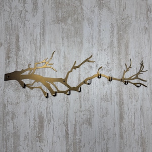 May include: A gold-colored metal wall-mounted coat rack shaped like a tree branch. The rack has multiple hooks for hanging items. The design features a branch with several smaller branches extending outwards, creating a natural and decorative look.