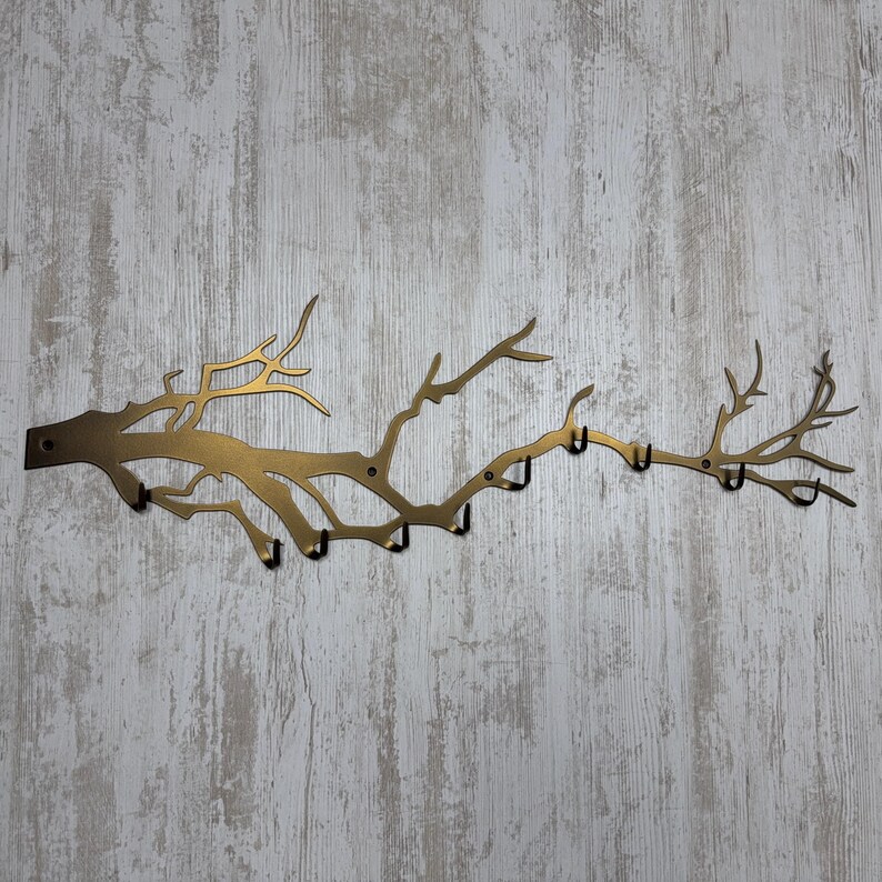 May include: A gold-colored metal wall-mounted coat rack shaped like a tree branch. The rack has multiple hooks for hanging items. The design features a branch with several smaller branches extending outwards, creating a natural and decorative look.