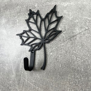 May include: Black metal wall hook shaped like a maple leaf. The hook is designed to hang items on a wall.