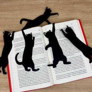 Pu&ograve; includere: Cinque segnalibri neri a forma di gatto sono posizionati su un libro aperto. I segnalibri sono realizzati in un materiale nero sottile e hanno la forma di gatti in varie pose. Il libro &egrave; aperto a una pagina con testo in una lingua straniera.