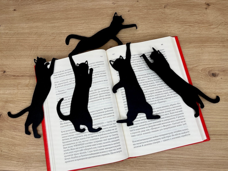 Pu&ograve; includere: Cinque segnalibri neri a forma di gatto sono posizionati su un libro aperto. I segnalibri sono realizzati in un materiale nero sottile e hanno la forma di gatti in varie pose. Il libro &egrave; aperto a una pagina con testo in una lingua straniera.