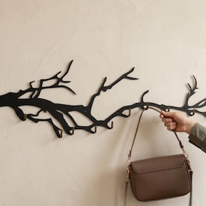 May include: Black metal wall-mounted coat rack shaped like a tree branch. The rack has multiple hooks for hanging items. A brown purse is hanging from one of the hooks. The rack is mounted on a light beige wall.