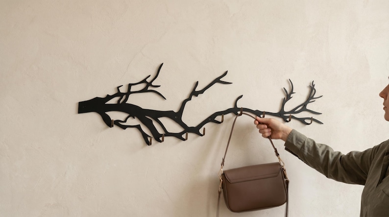 May include: Black metal wall-mounted coat rack shaped like a tree branch. The rack has multiple hooks for hanging items. A brown purse is hanging from one of the hooks. The rack is mounted on a light beige wall.