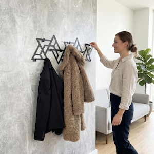 May include: A black geometric wall-mounted coat rack with three sections, holding a black jacket and a tan faux fur coat. The rack is mounted on a textured gray wall. A person is reaching for keys on the rack.