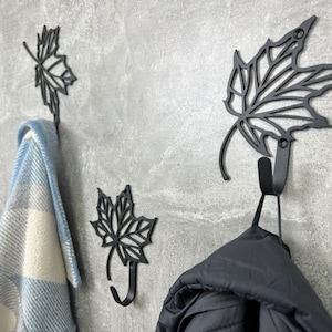 May include: Three black metal leaf-shaped hooks mounted on a gray wall. A blue and white plaid blanket hangs on the left hook, and a black jacket hangs on the right hook.