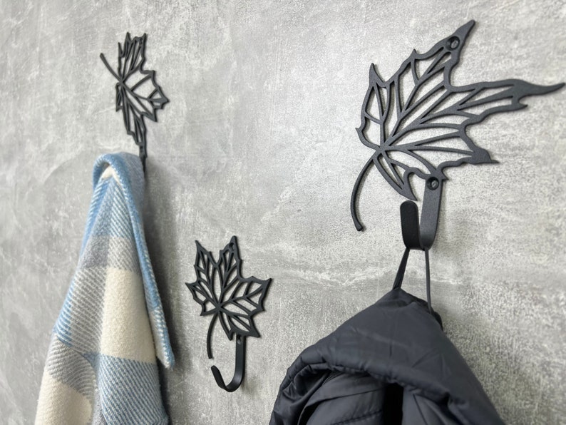 May include: Three black metal leaf-shaped hooks mounted on a gray wall. A blue and white plaid blanket hangs on the left hook, and a black jacket hangs on the right hook.