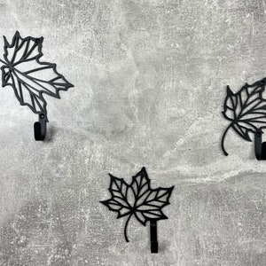 May include: Three black metal leaf-shaped hooks on a gray textured wall. Each hook is shaped like a maple leaf with a rounded hook at the bottom.
